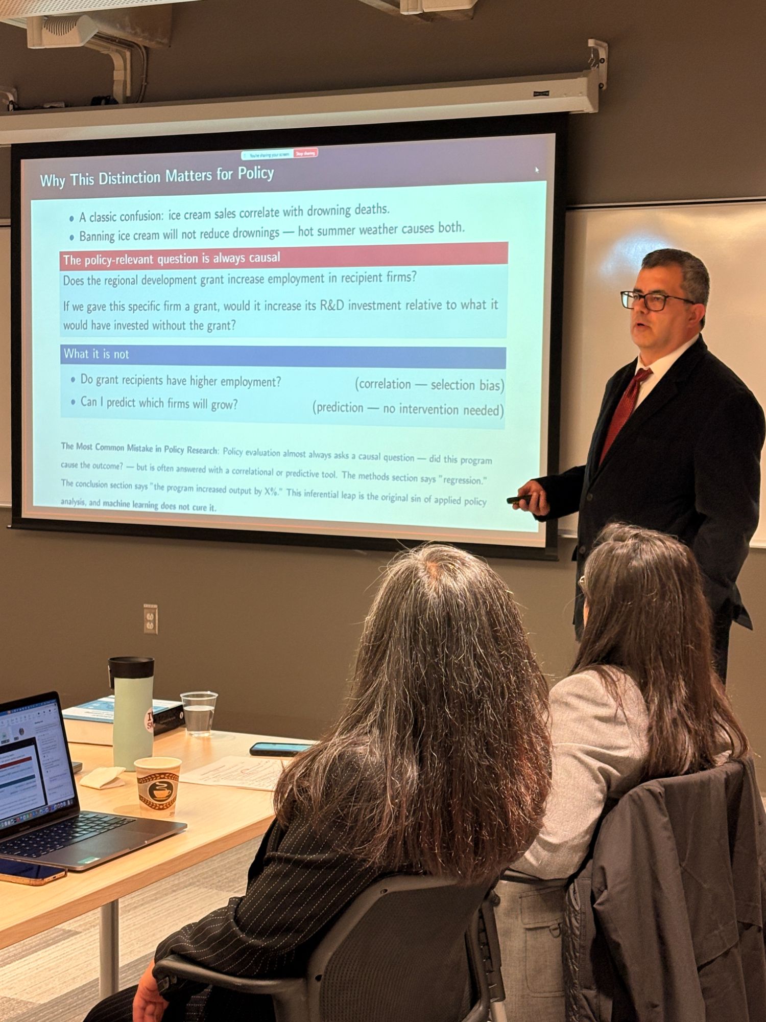Keynote speaker presents a slide titled 'Why This Distinction Matters for Policy', contrasting causal questions with correlation and prediction.