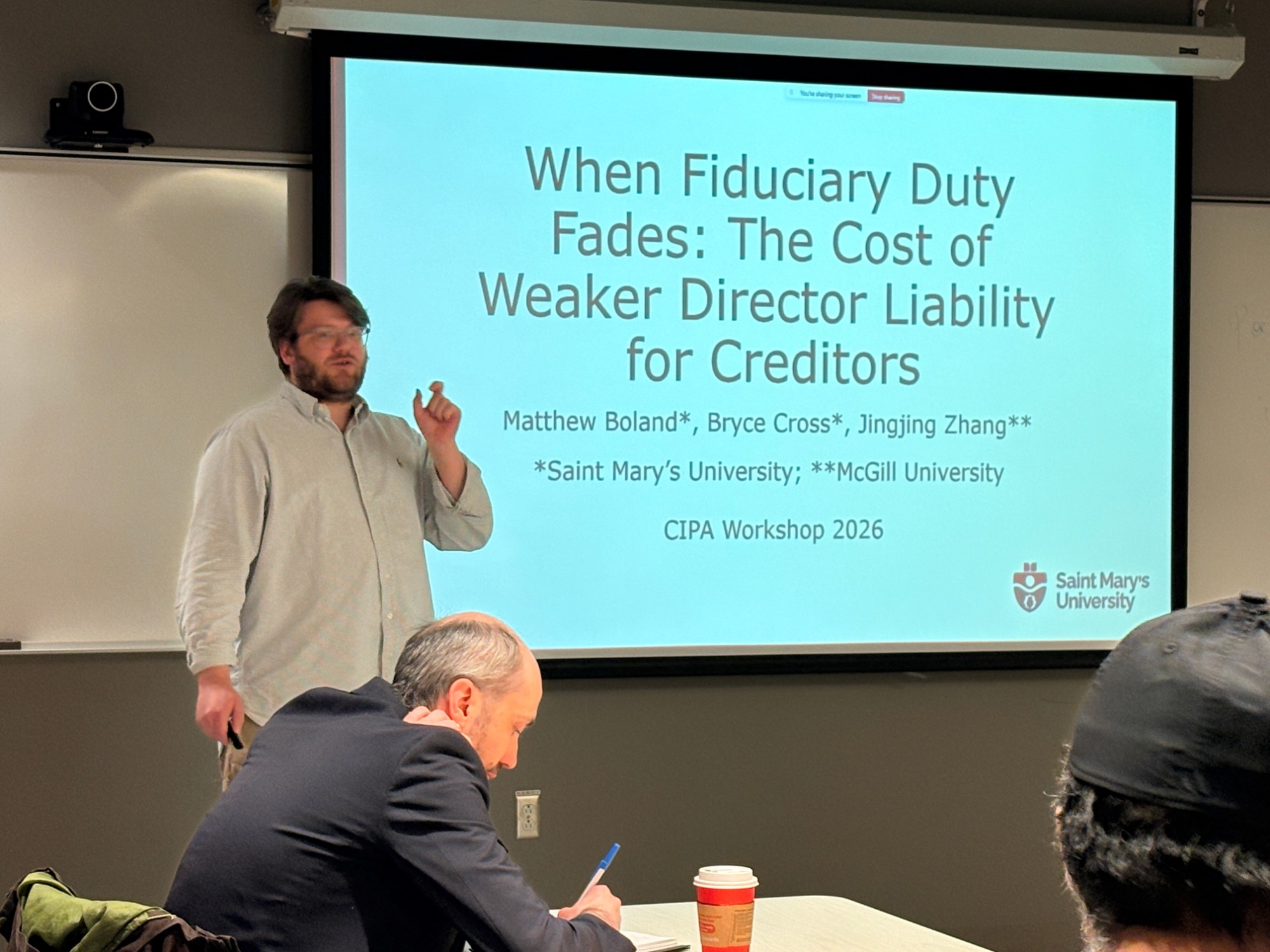A presenter stands beside a slide titled 'When Fiduciary Duty Fades: The Cost of Weaker Director Liability for Creditors' while a participant takes notes.